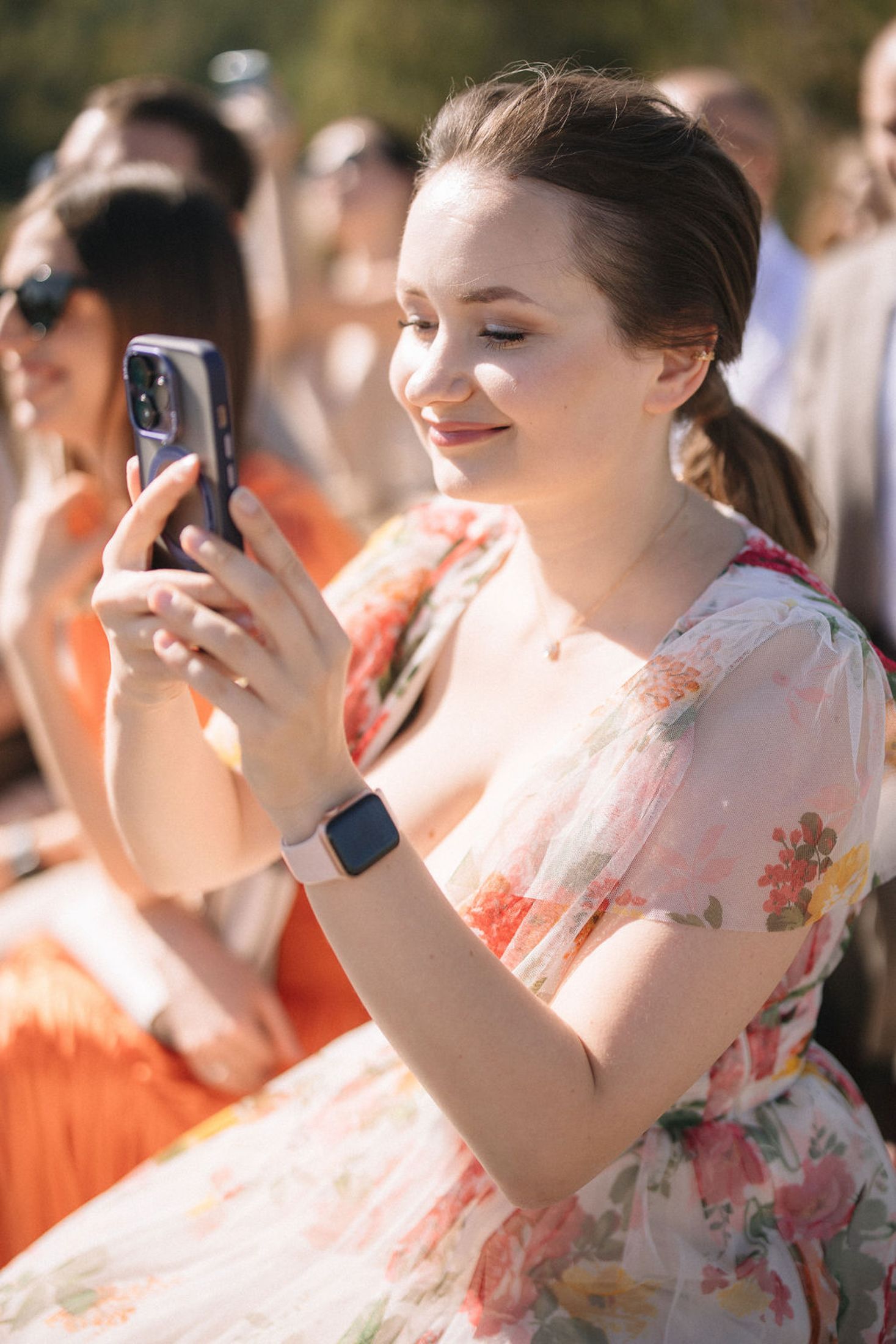 A person in a floral dress holds up a smartphone while wearing a smartwatch at an outdoor gathering.