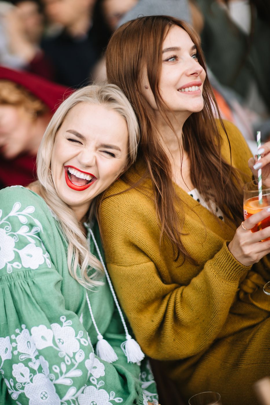 Two friends laughing together while enjoying drinks, one wearing a mint green floral top and another in mustard yellow.