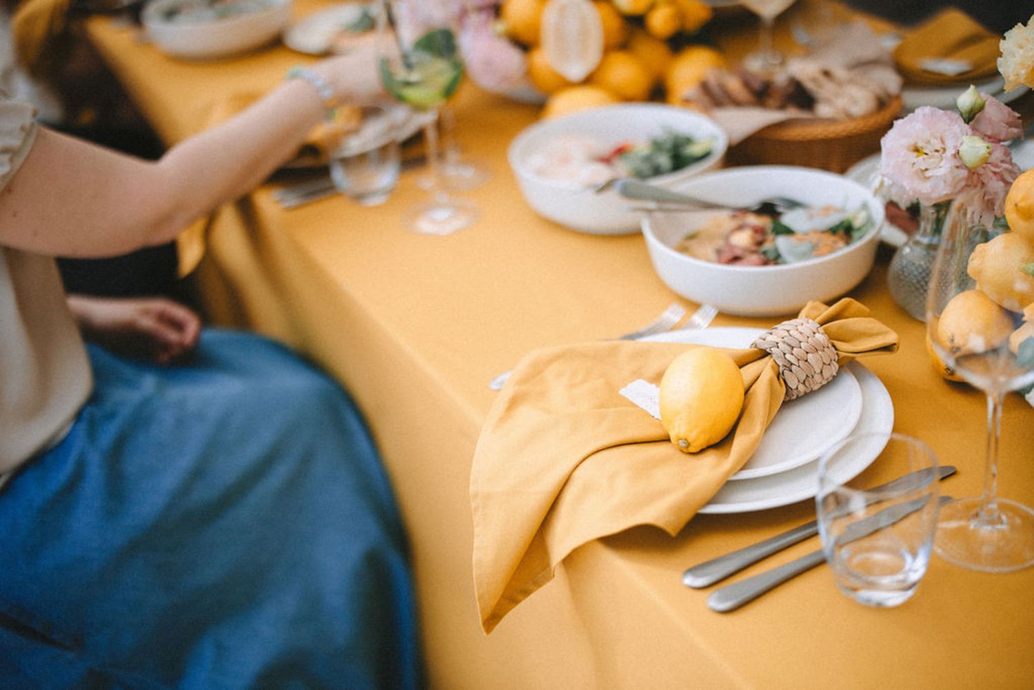 A table set with yellow tablecloth and white plates for a dining event with hands visible reaching across.