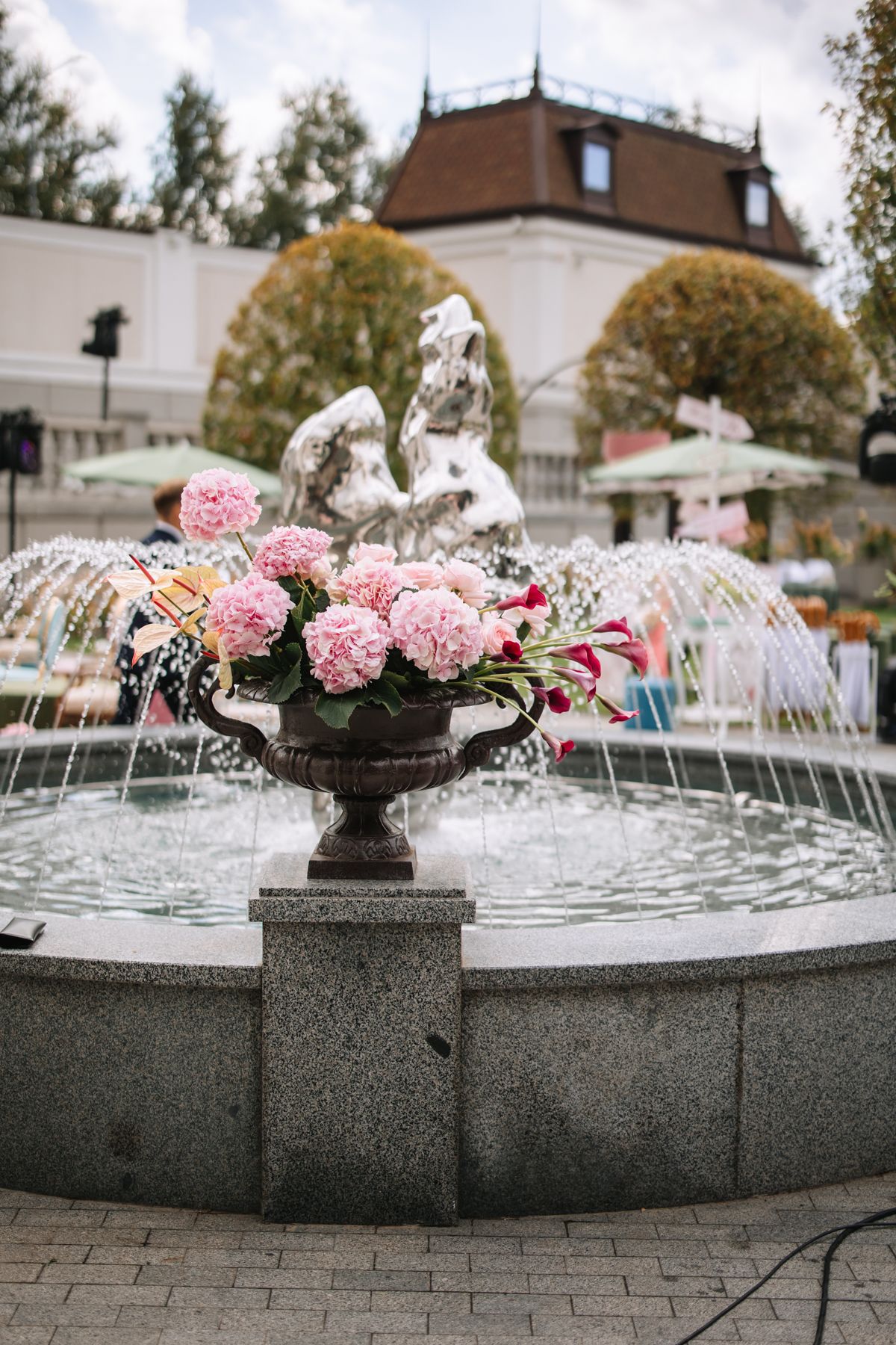 A decorative fountain with pink peonies and cherub statues sprays water in an elegant garden setting.