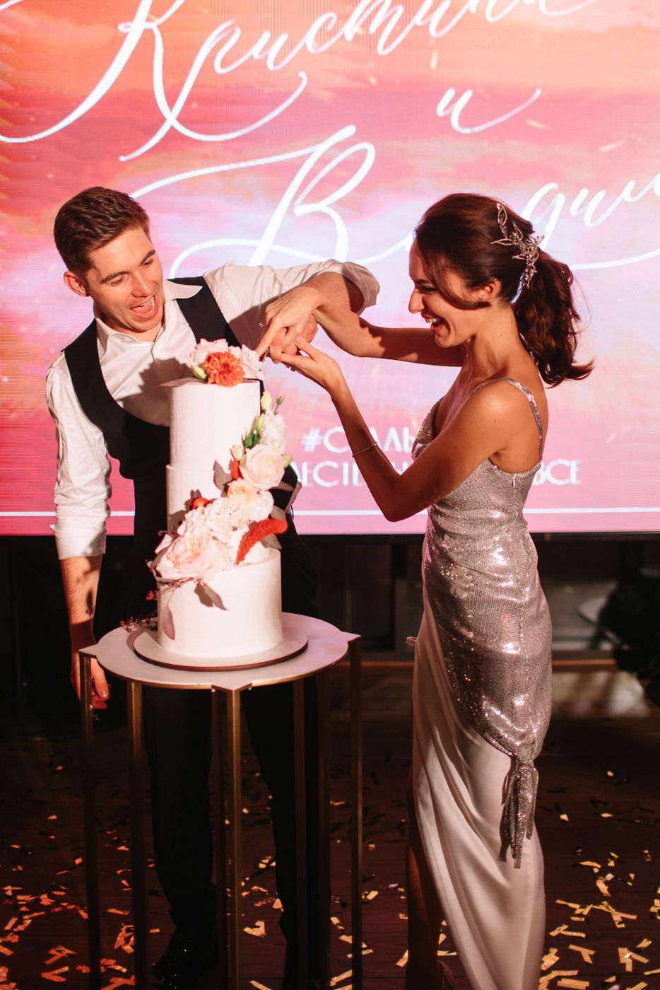 Wedding couple cutting and feeding each other cake with orange backdrop lighting.