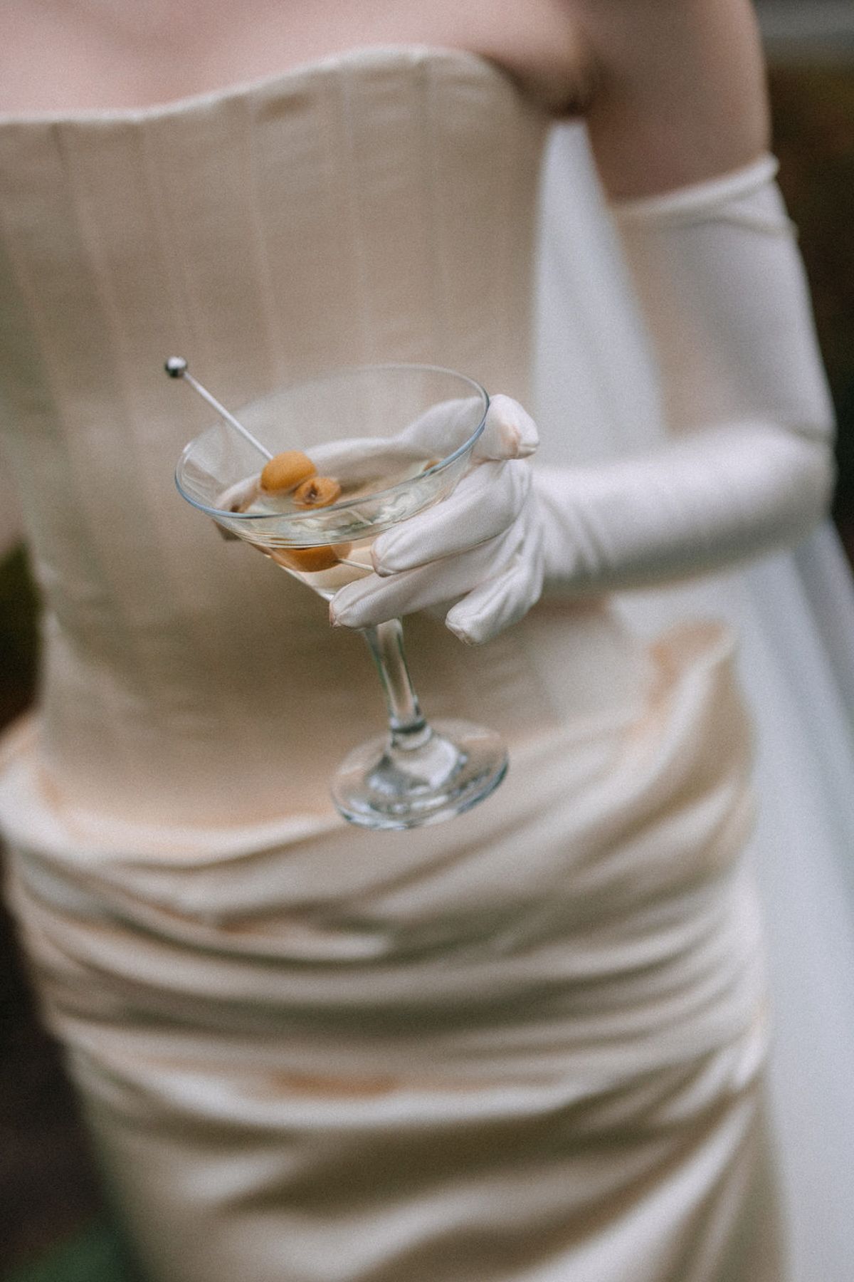 A gloved hand holds a martini glass with an olive garnish against a backdrop of an elegant cream-colored dress.