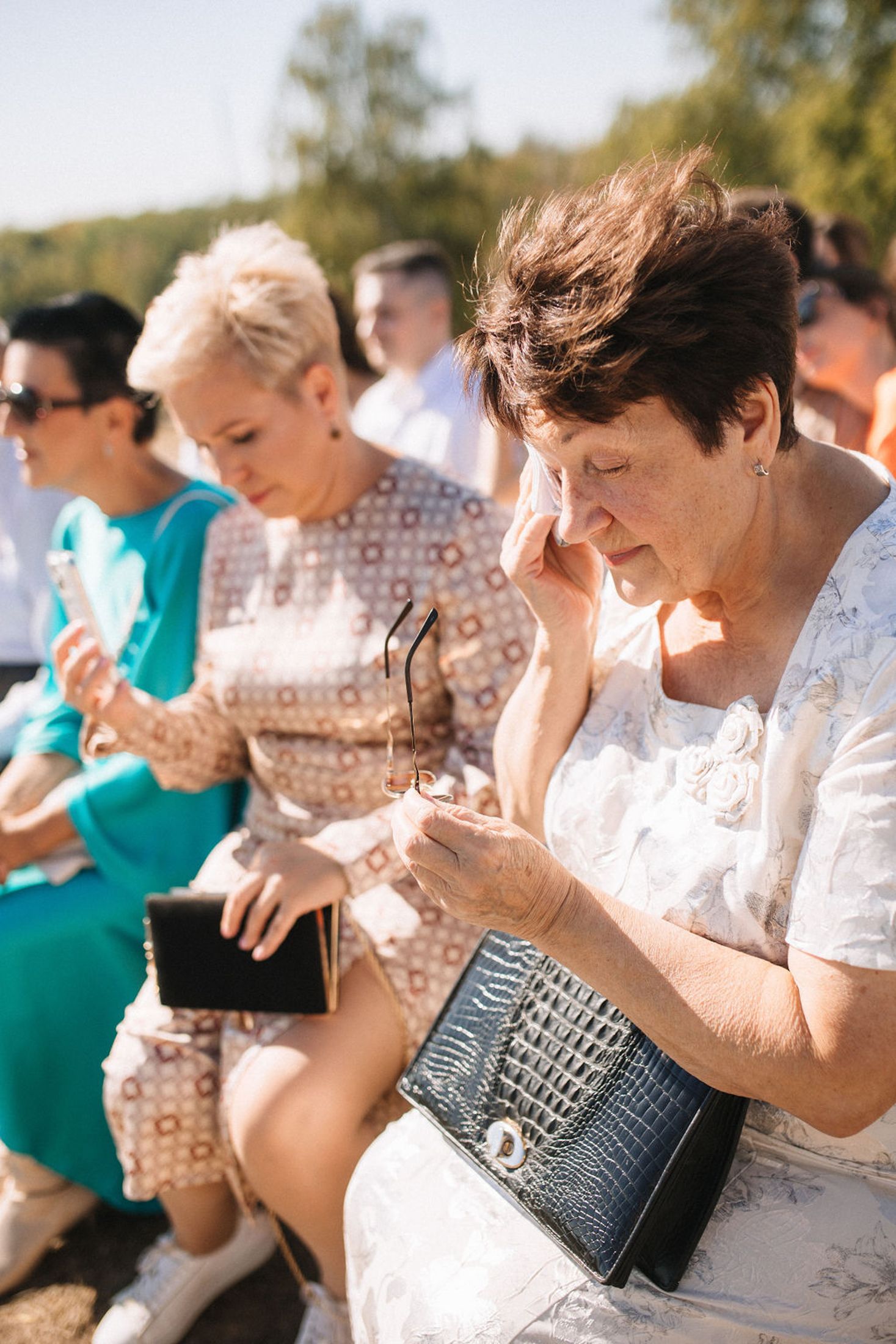 People in summer attire check their phones while sitting outdoors at a sunny event.