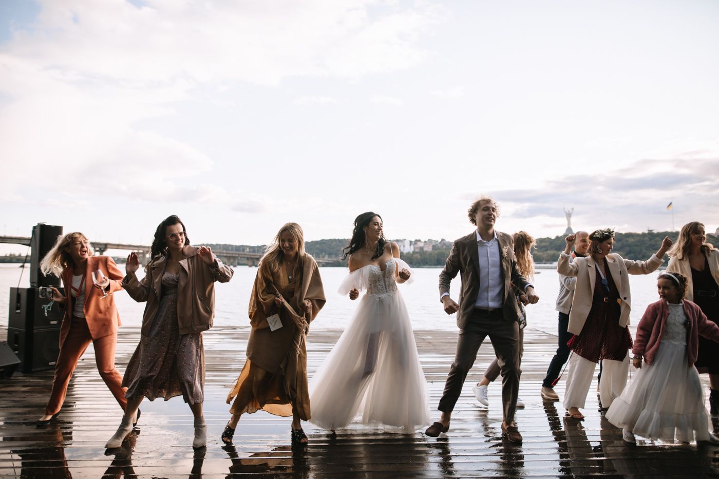 Wedding party members stand together on a beach pier during sunset creating silhouettes against the orange sky.