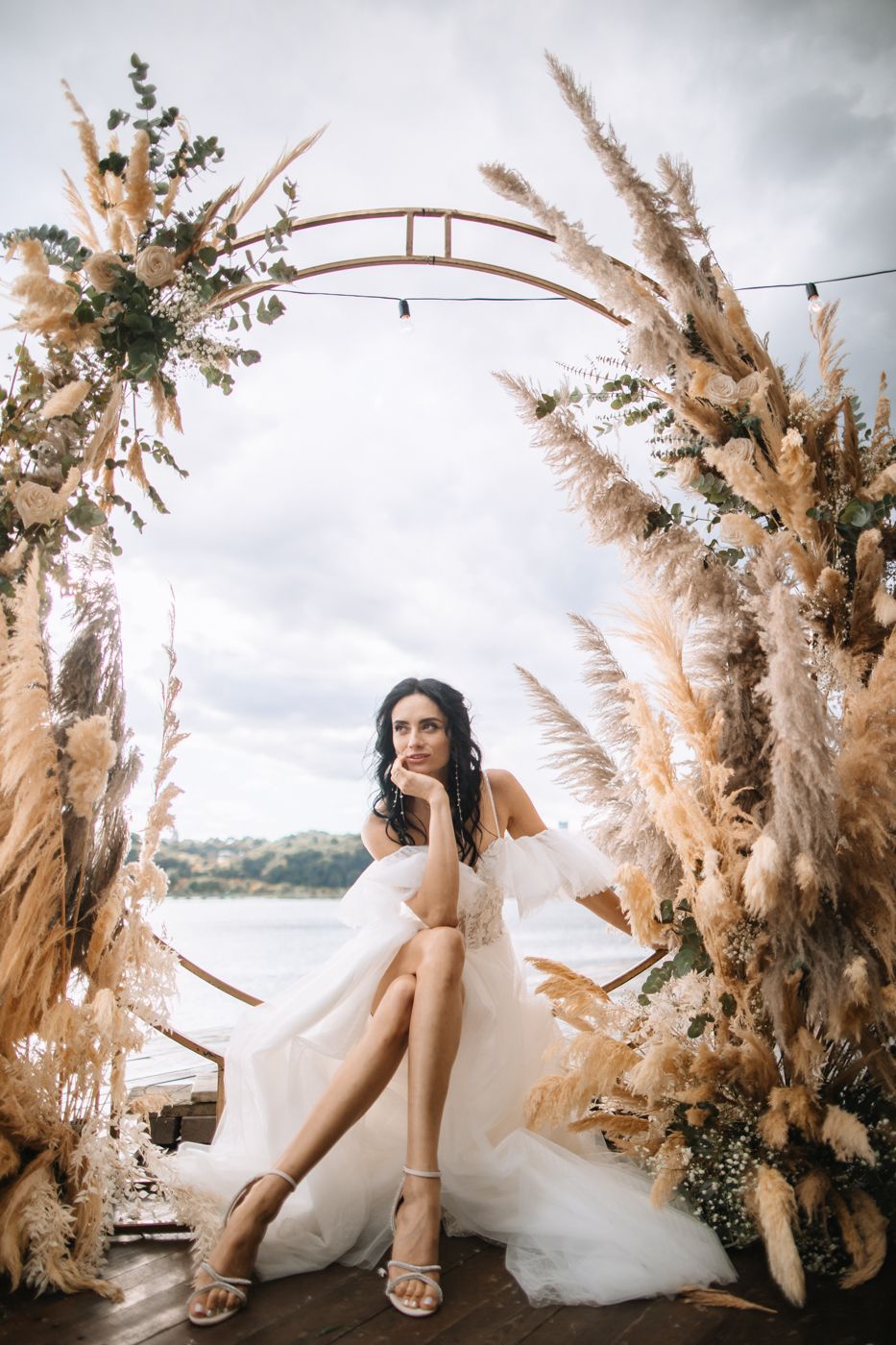 Elegant wedding details featuring pampas grass floral arrangements and a flowing white bridal gown against a cloudy sky.