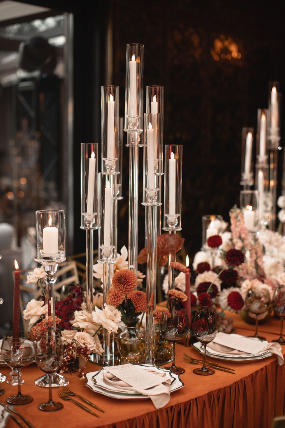 Elegant table setting with tall glass candleholders and autumn-colored floral arrangements.