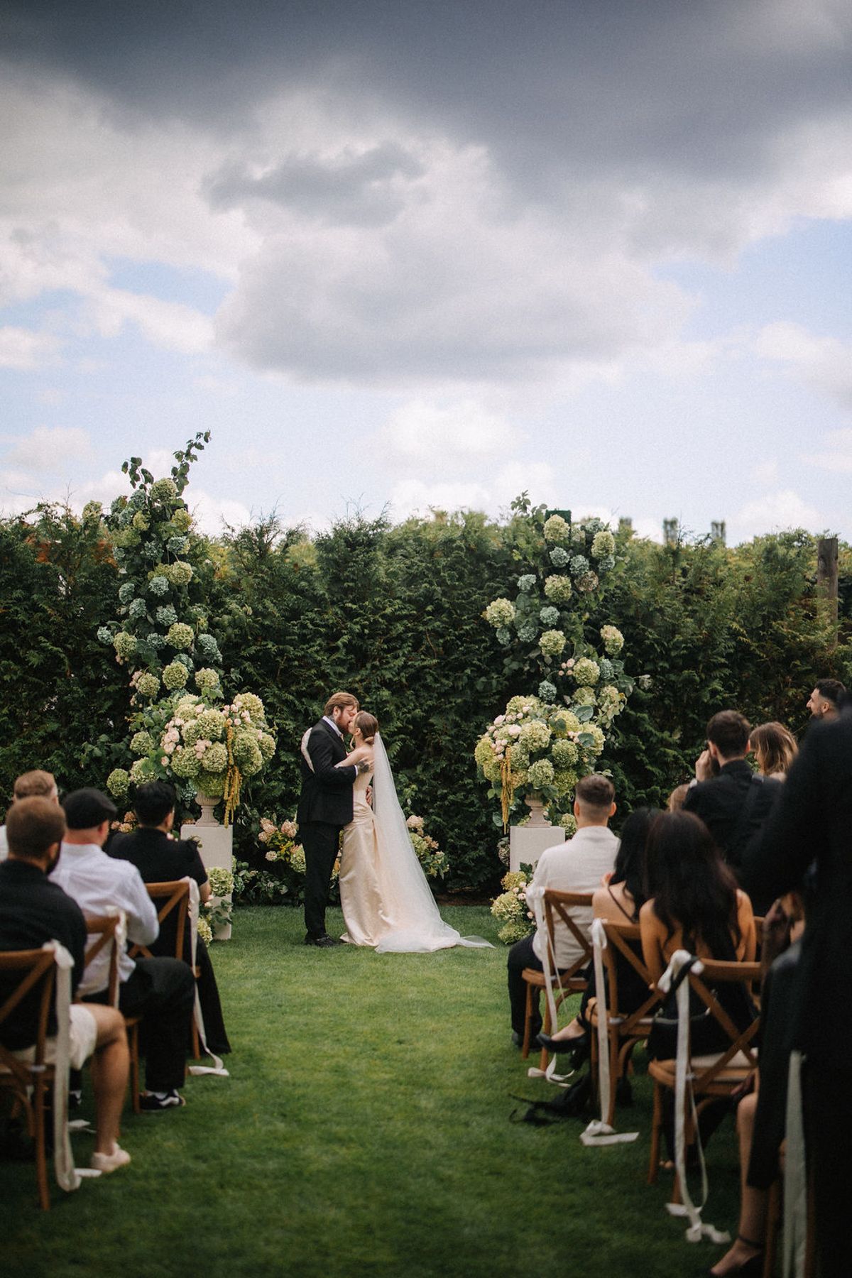 Panoramic view of an outdoor wedding ceremony on a lush green lawn with white floral arrangements and wooden chairs.