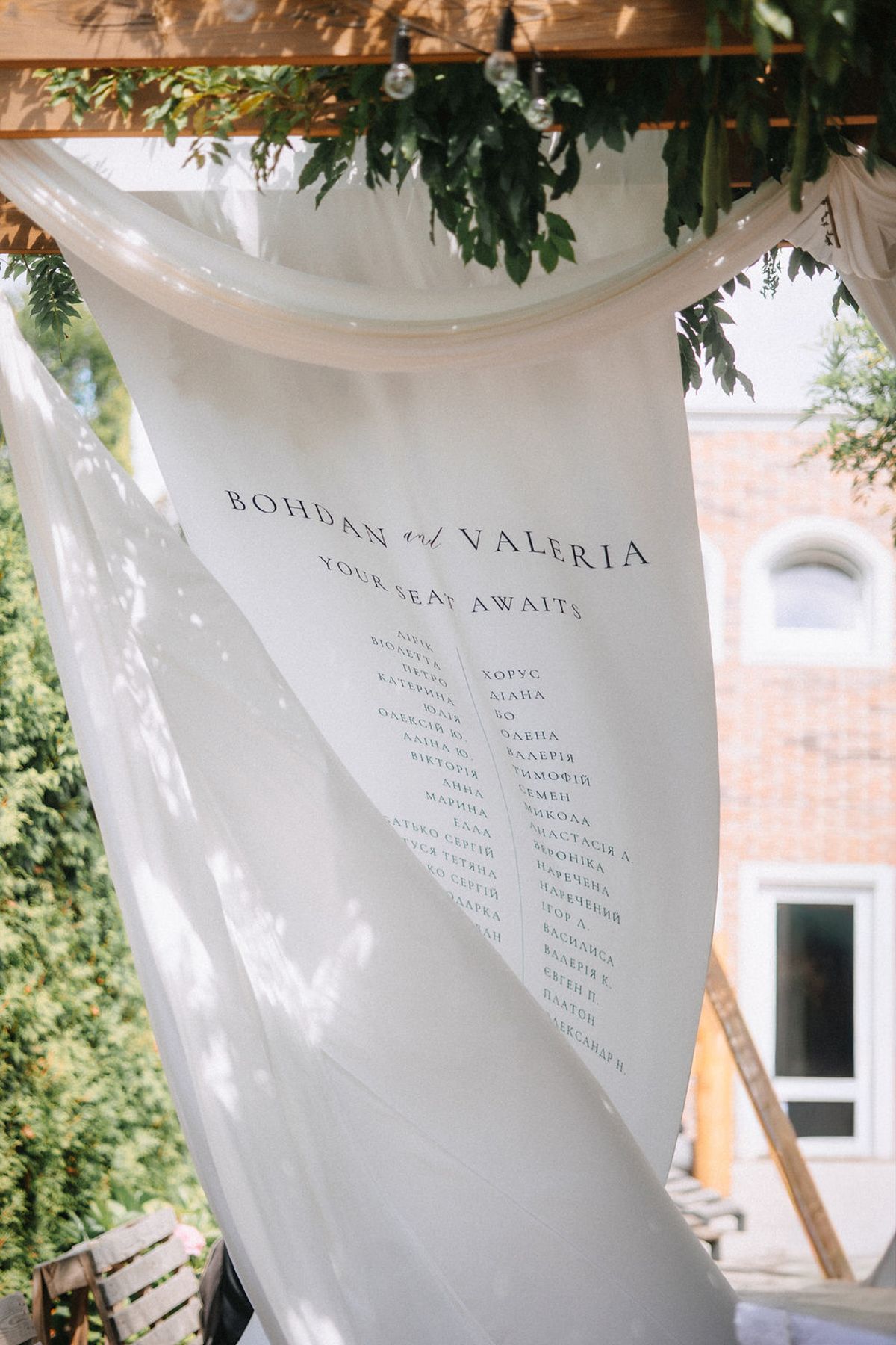 Translucent white wedding seating chart banners with names flutter gently in the breeze against a green foliage backdrop.