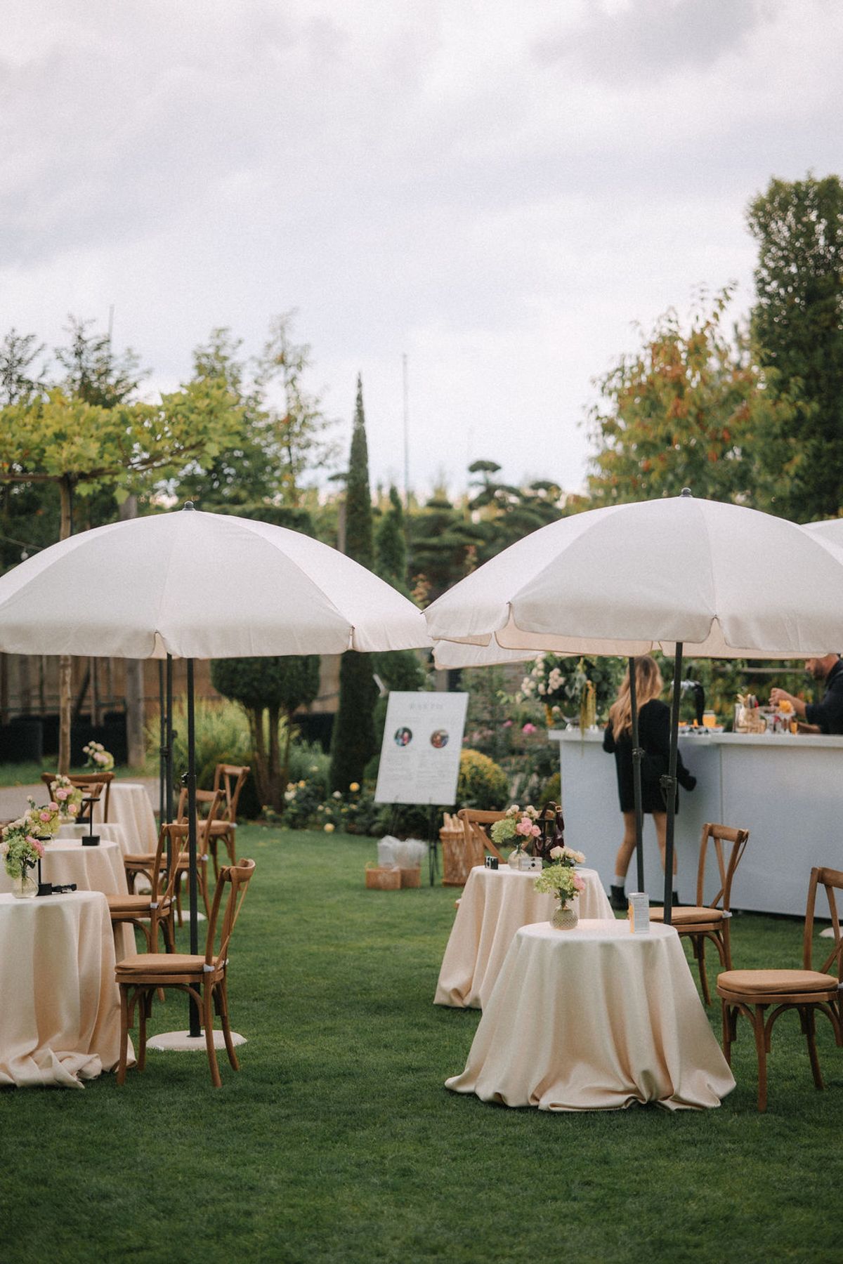 White umbrellas and round tables with cream linens create an elegant outdoor reception setup on a lush green lawn.