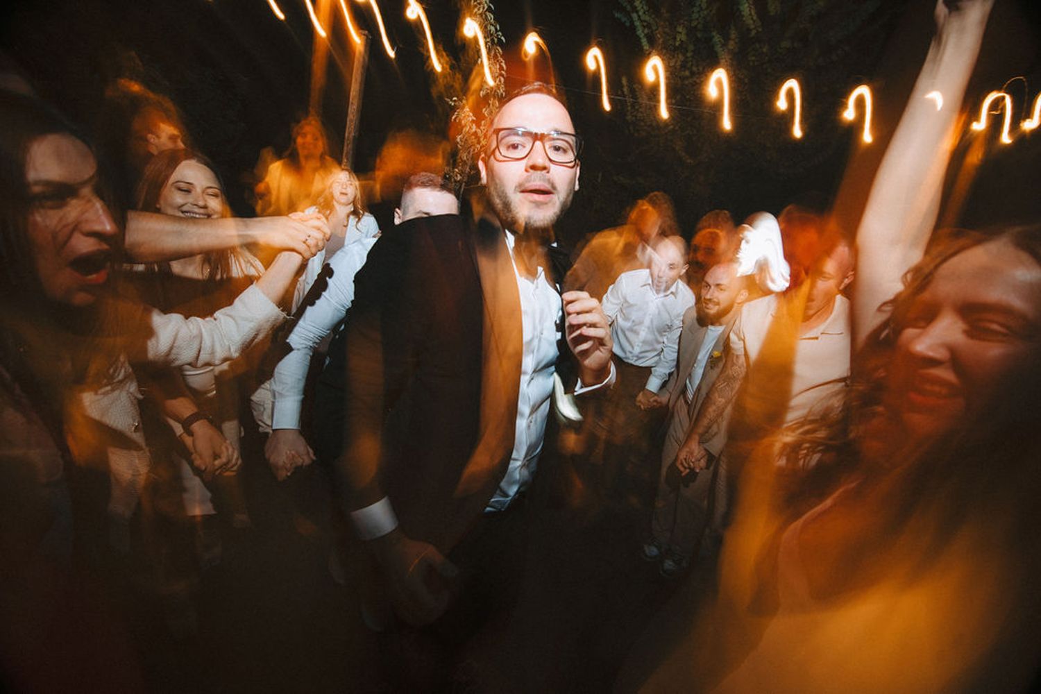 A lively wedding reception dance party with string lights overhead and guests celebrating on a crowded dance floor.