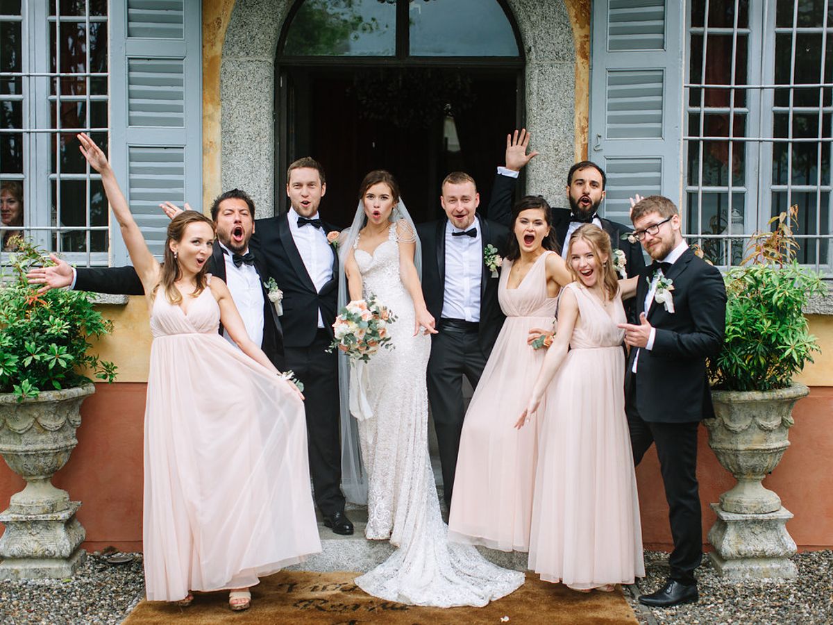 A wedding party poses in front of a blue mansion with bridesmaids in pink dresses and groomsmen in dark suits.