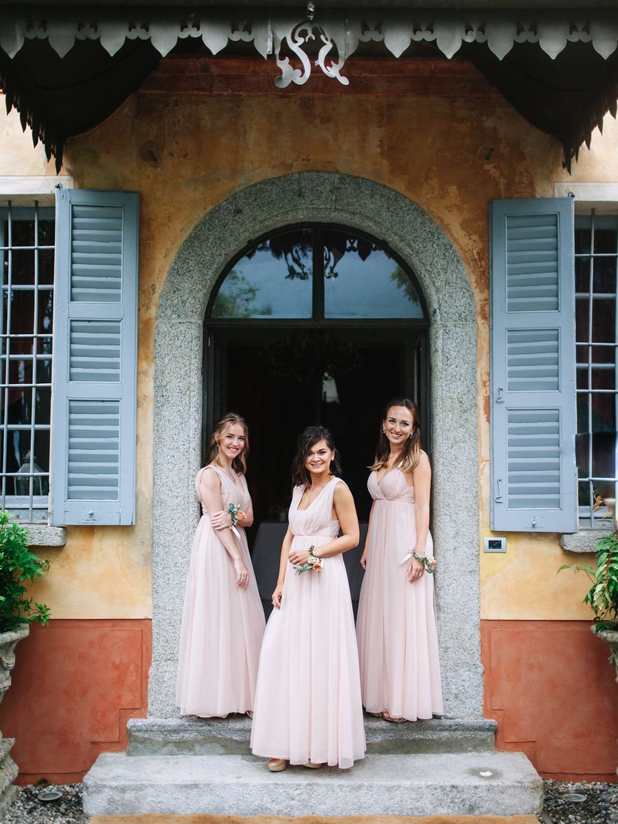 Three people in matching blush pink bridesmaid dresses pose in an elegant arched doorway with blue shutters.