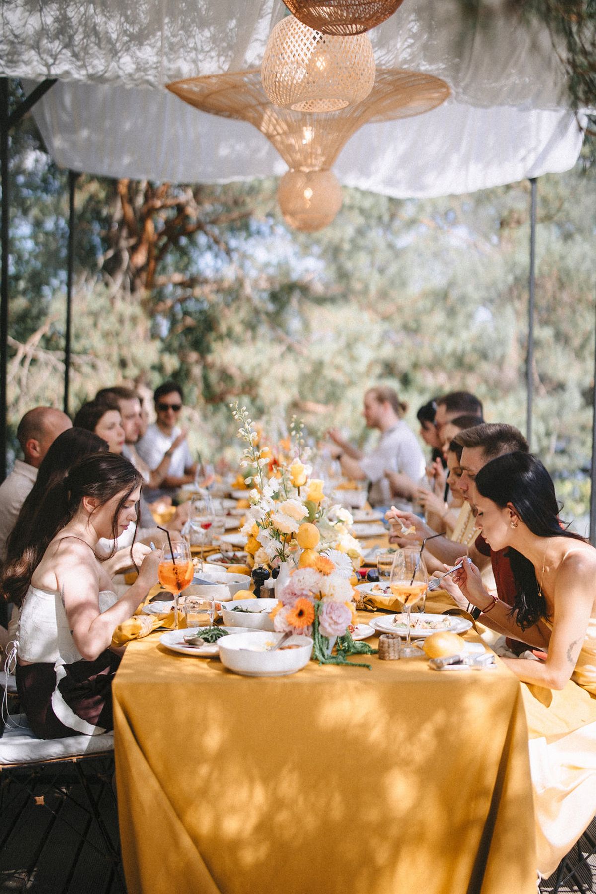 Wedding guests gather around elegant tables with yellow tablecloths and floral centerpieces under white canopy outdoors.