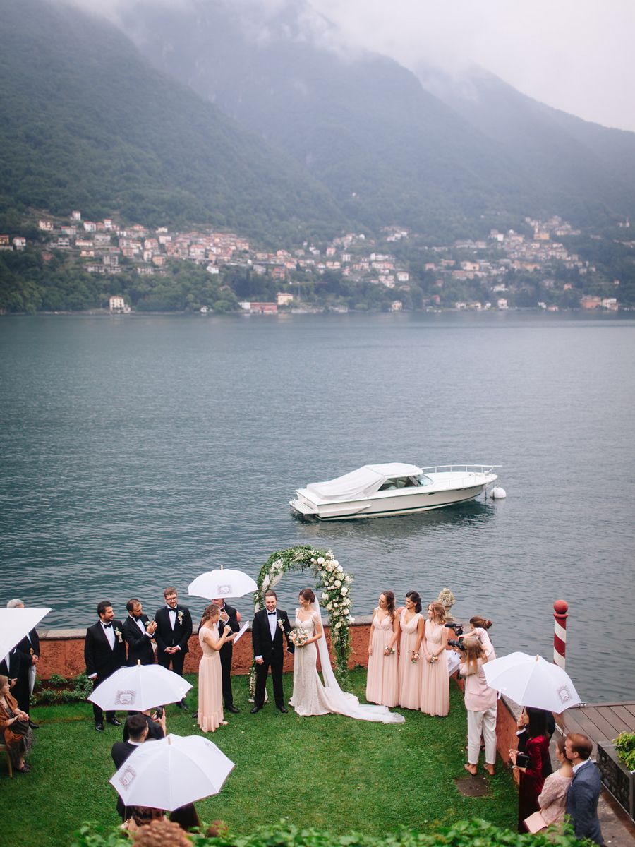 Scenic lakeside wedding ceremony in Italy with mountains and small village in background, boats on tranquil water.
