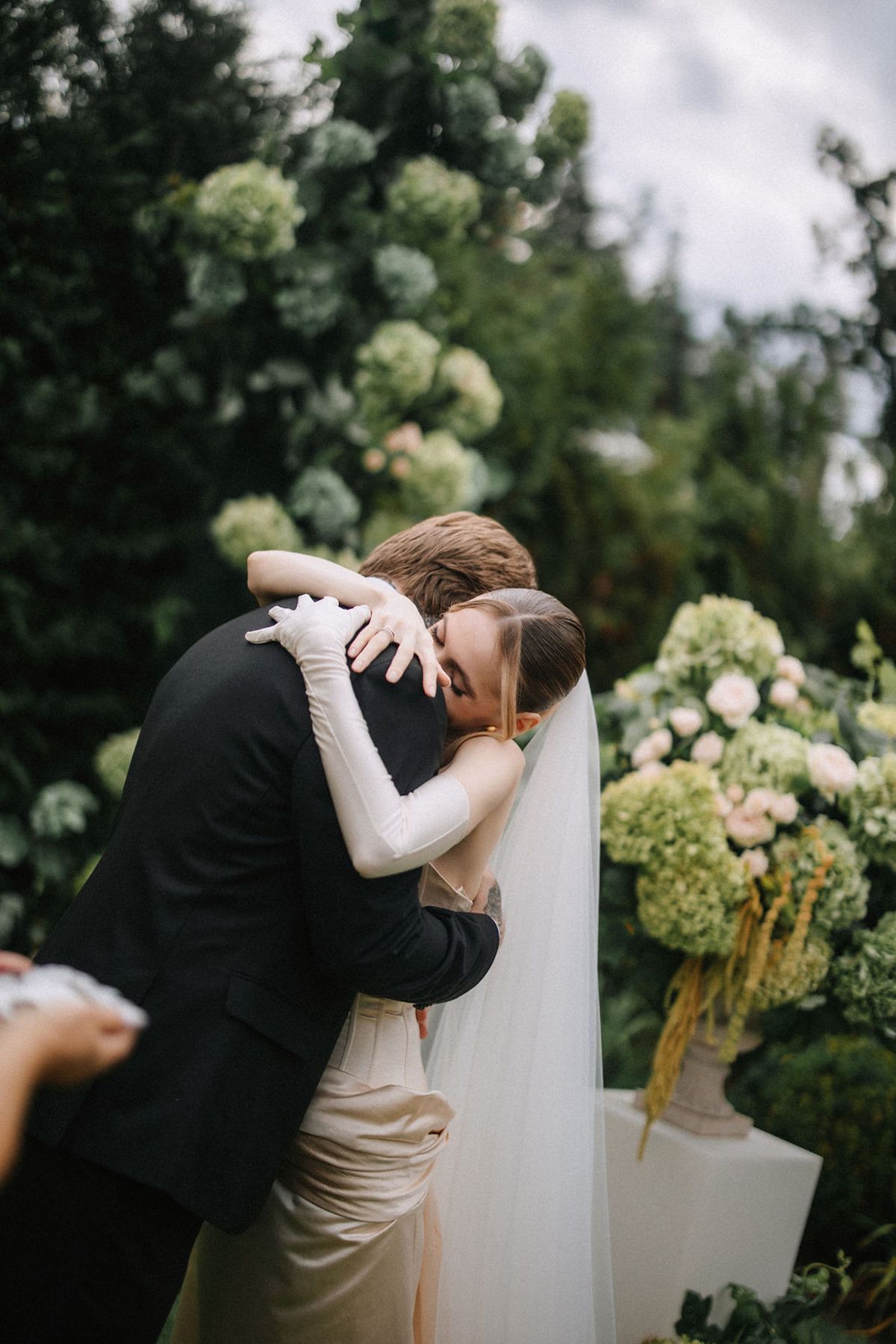 A couple shares a romantic kiss during their outdoor garden wedding ceremony surrounded by white hydrangea flowers.