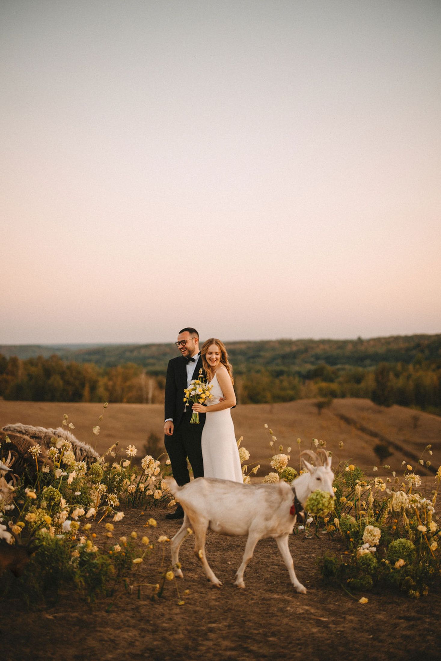 A couple in wedding attire stands on a hilltop at sunset with a white deer walking beside them among yellow wildflowers.