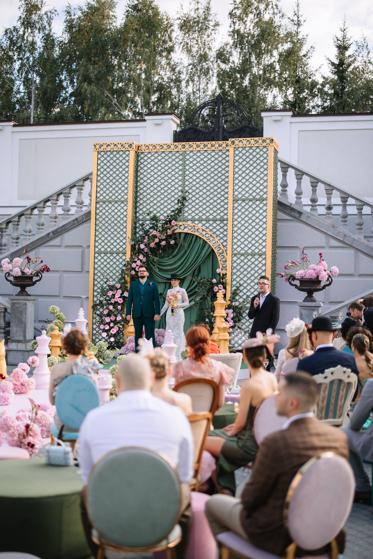 Wedding ceremony guests seated outdoors watching couple exchange vows under a decorative lattice arch with floral accents.