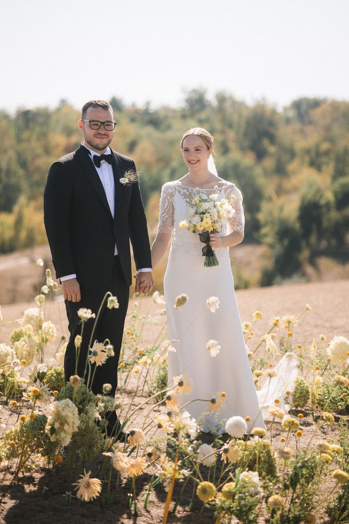 A couple in wedding attire stands together in a field of blooming yellow wildflowers on a sunny autumn day.