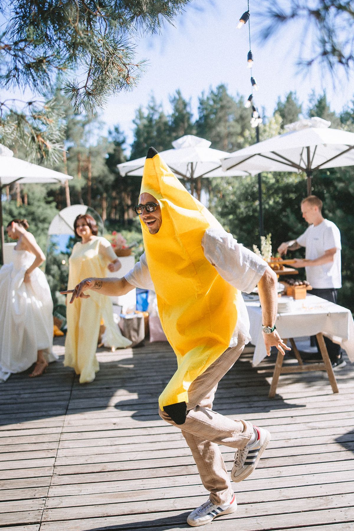 Someone in a bright yellow banana costume dances energetically on a wooden deck with white umbrellas and trees in background.