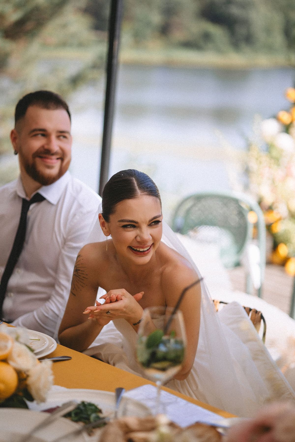 Wedding reception guests laugh and share joyful moments at a sunny outdoor table setting by the water.