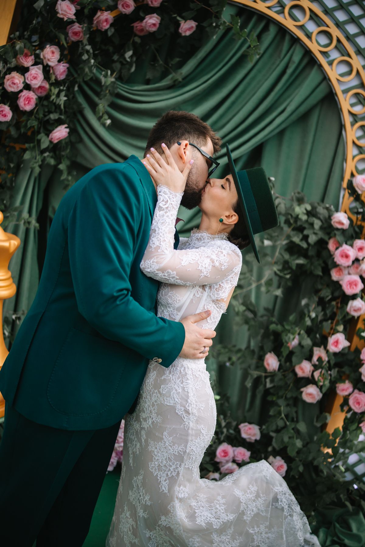 Romantic wedding couple embraces under floral arch with pink roses and emerald green decor.