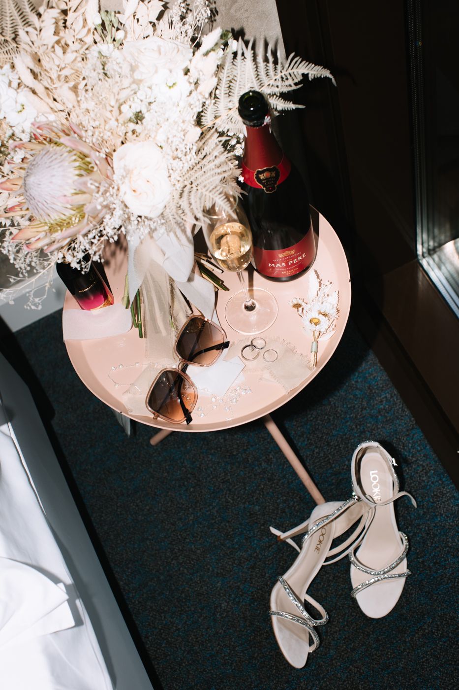 A pink side table displays champagne, sunglasses, and silver sandals with a white floral arrangement.