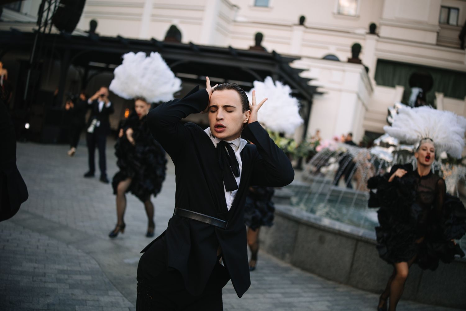 A person in a black tuxedo dances energetically with white pom poms in an outdoor plaza.