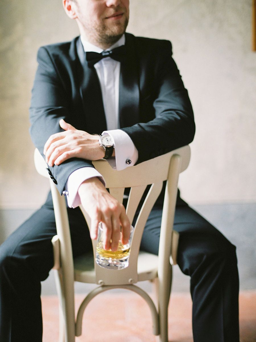 A person in a dark suit sits on a wooden chair holding a glass of amber liquid against a light neutral background.