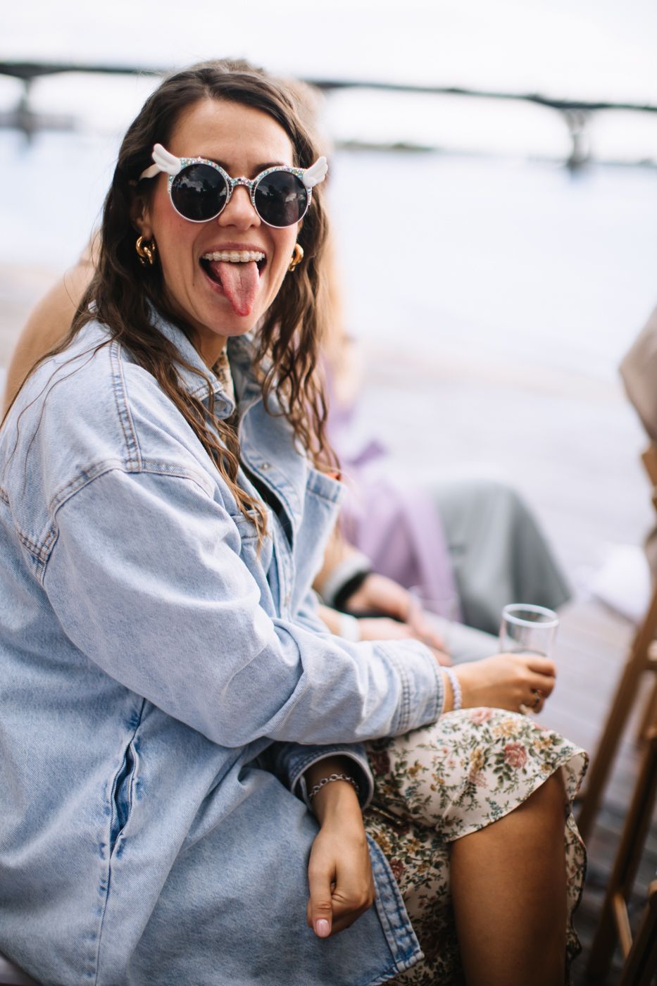 Woman in denim shirt laughing and sticking out tongue while sitting outdoors.