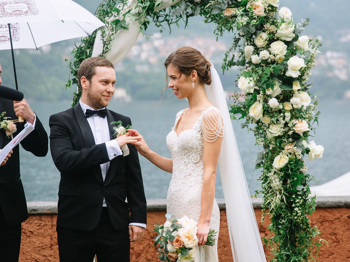 A romantic outdoor wedding ceremony under a floral arch with white roses against a misty mountain backdrop.