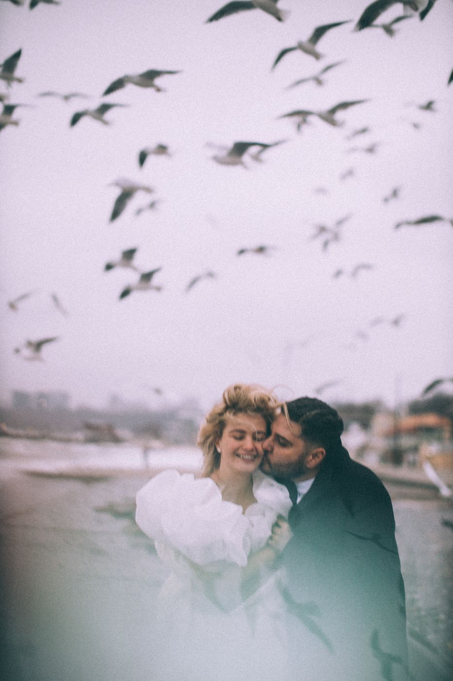 Romantic couple embraces on beach with flock of birds flying above.