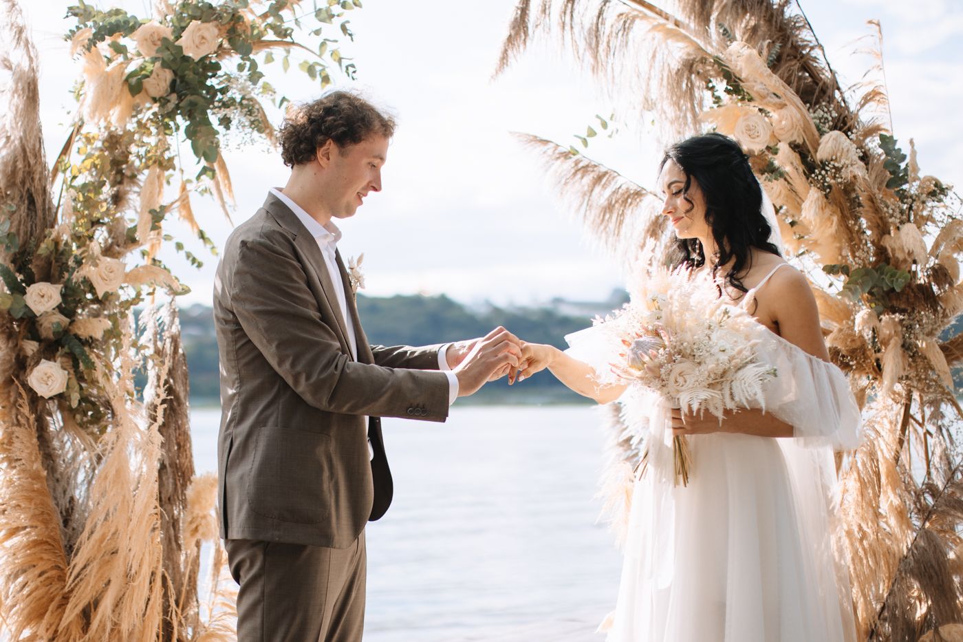 A couple exchanges vows under a floral arch with pampas grass and dried flowers overlooking a scenic lake view.