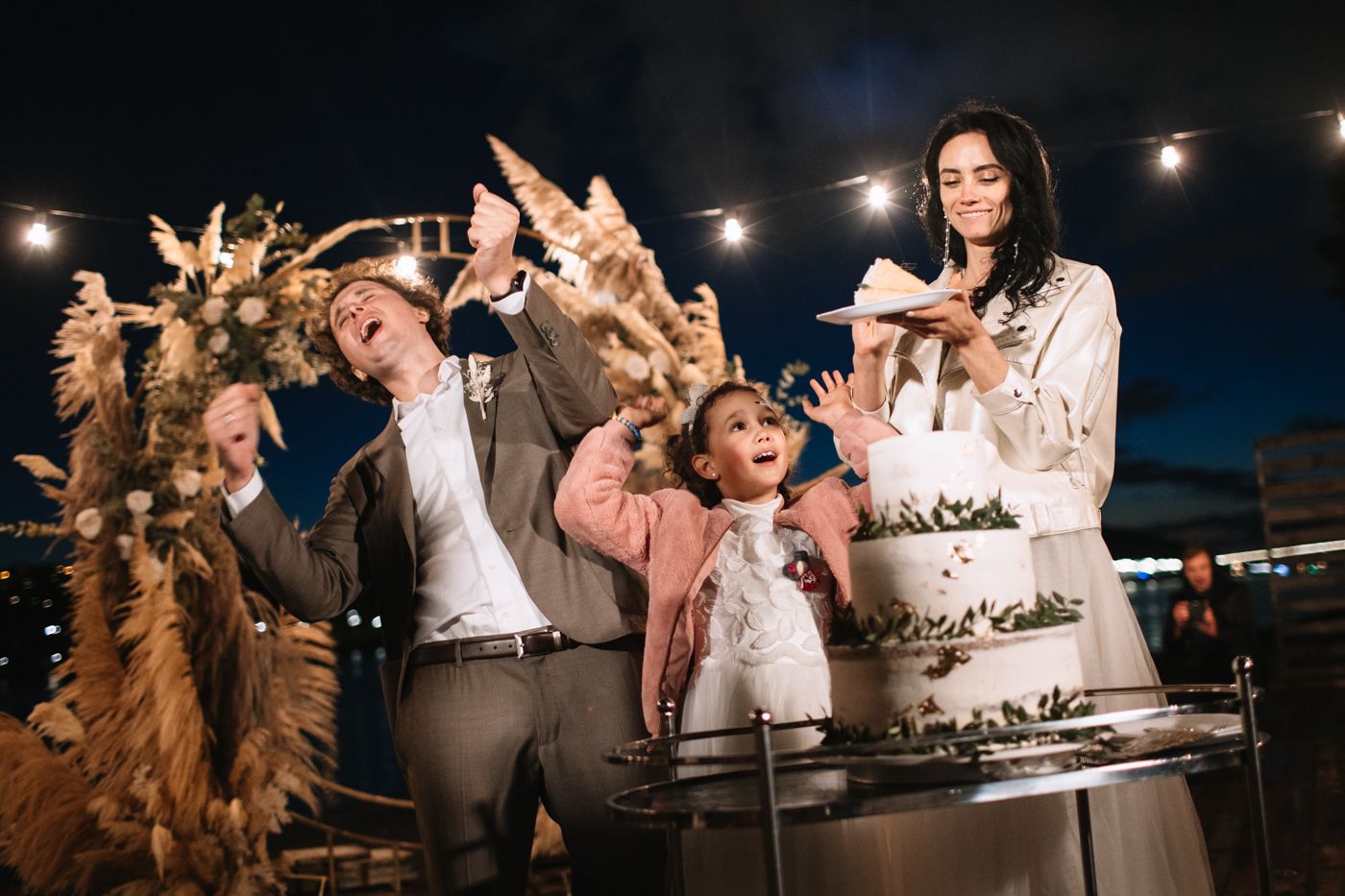 Wedding guests gather around a multi-tiered white cake at an outdoor evening reception as sparklers illuminate the celebration.