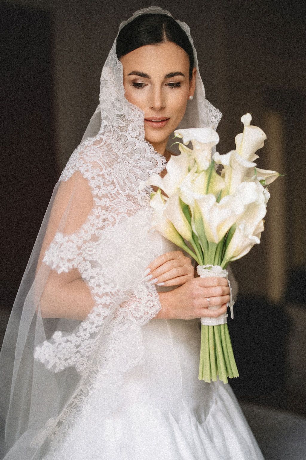 A bride in a white lace wedding dress holds a bouquet of white calla lilies and smiles while looking down at her flowers.