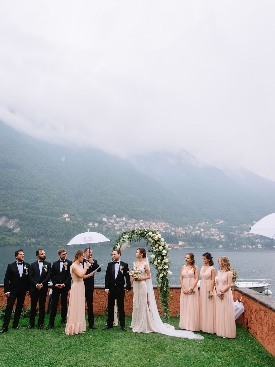 Wedding party poses on grassy hill overlooking scenic Lake Como in Italy with misty mountains in background.