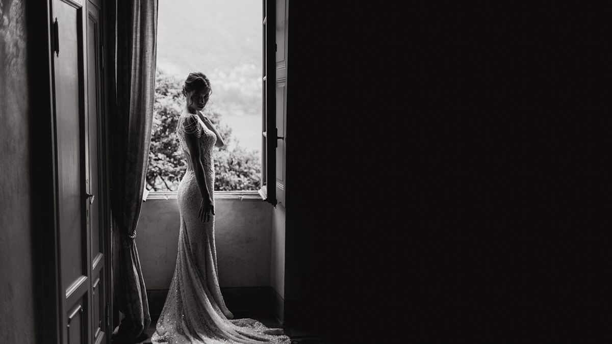 A silhouette of a bride in a fitted wedding dress stands by a window in dramatic black and white.