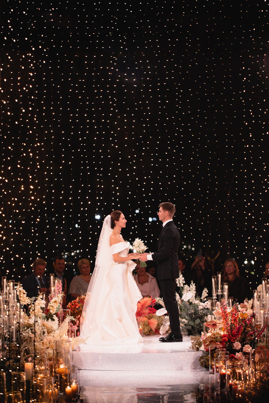 A wedding ceremony setup with fairy light backdrop and candlelit aisle decorations.