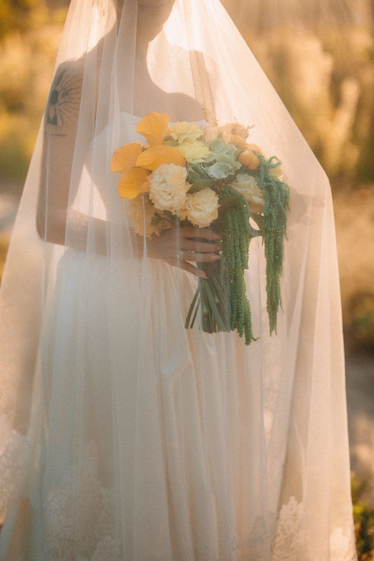 A delicate bridal bouquet with yellow and white flowers and cascading greenery seen through a sheer veil in golden light.
