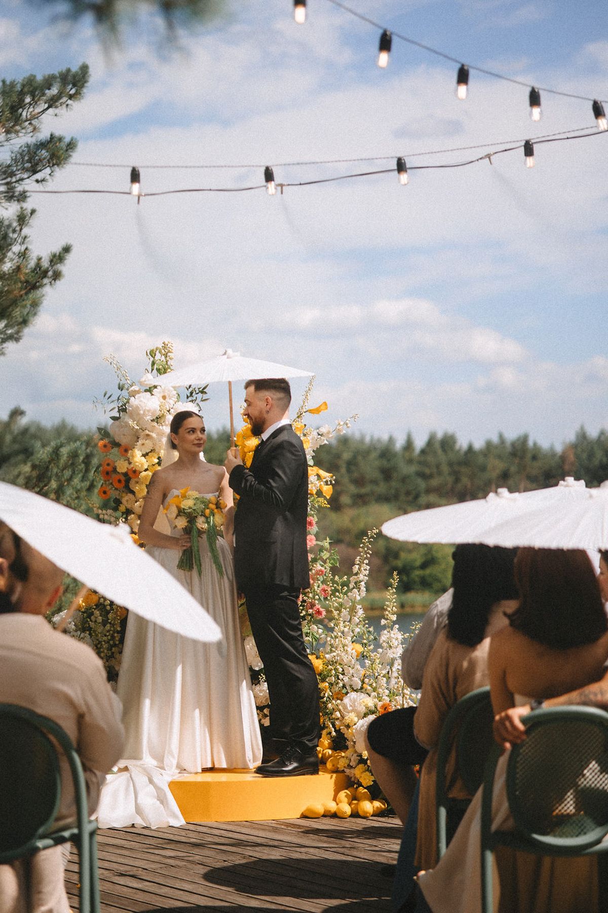 A romantic outdoor wedding ceremony on a wooden deck with string lights overhead and white parasols in the audience.