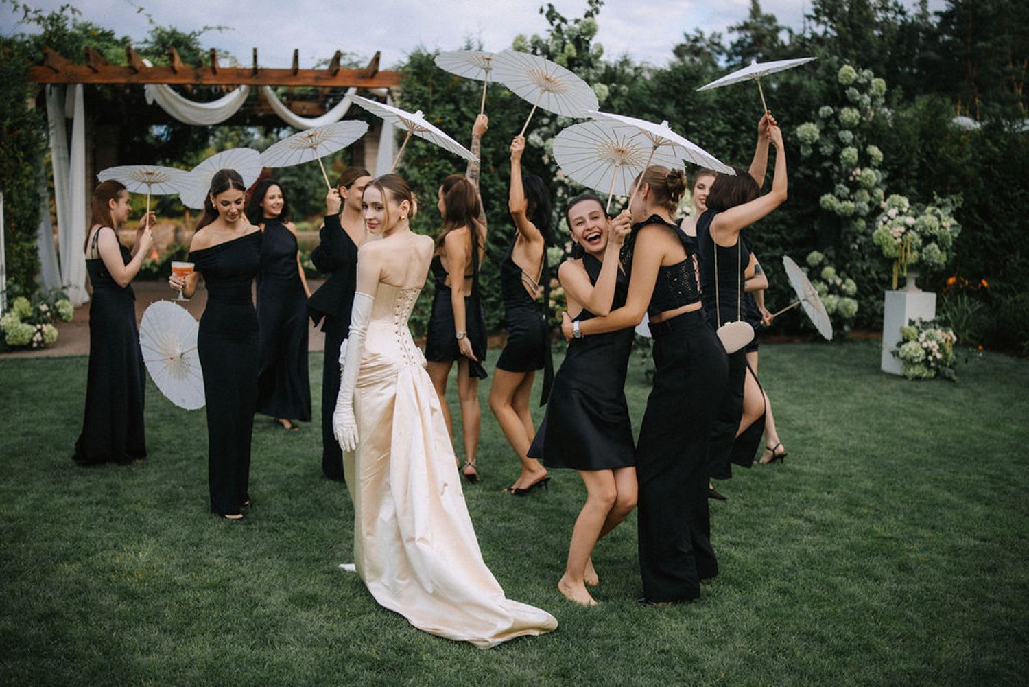 Wedding party celebrates on a lawn with bridesmaids in black dresses dancing under a decorated pergola at an outdoor reception.