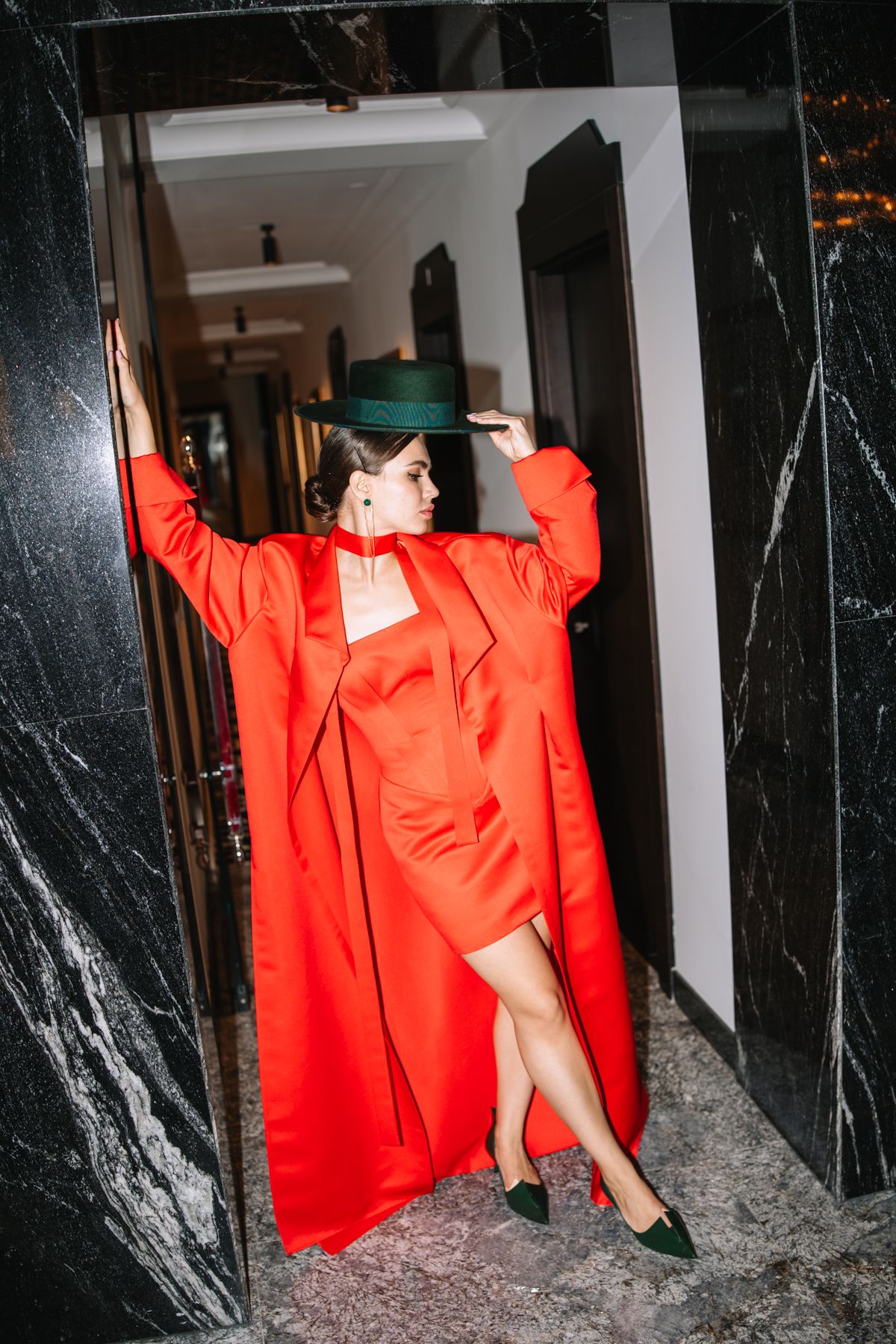 Person in bright red graduation gown and cap poses dramatically in doorway against dark marble walls.