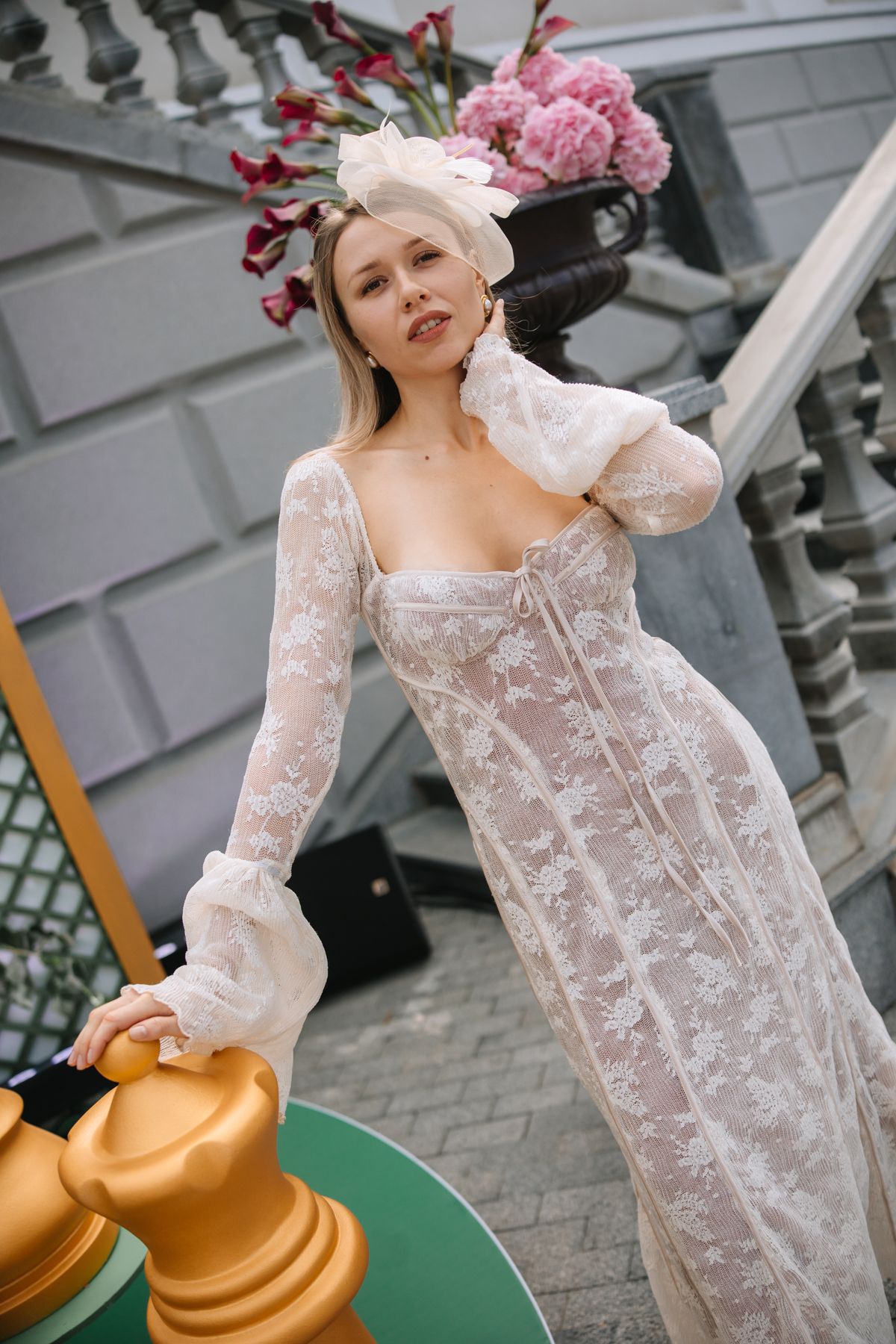 A person in a white lace dress poses with a golden decorative element against an industrial gray background.