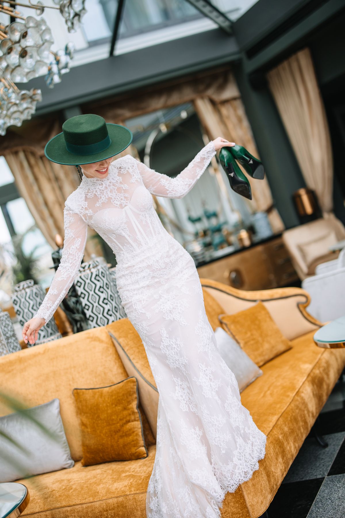 Graceful pose in vintage-style white lace gown and green hat against velvet couch in upscale room with chandelier.