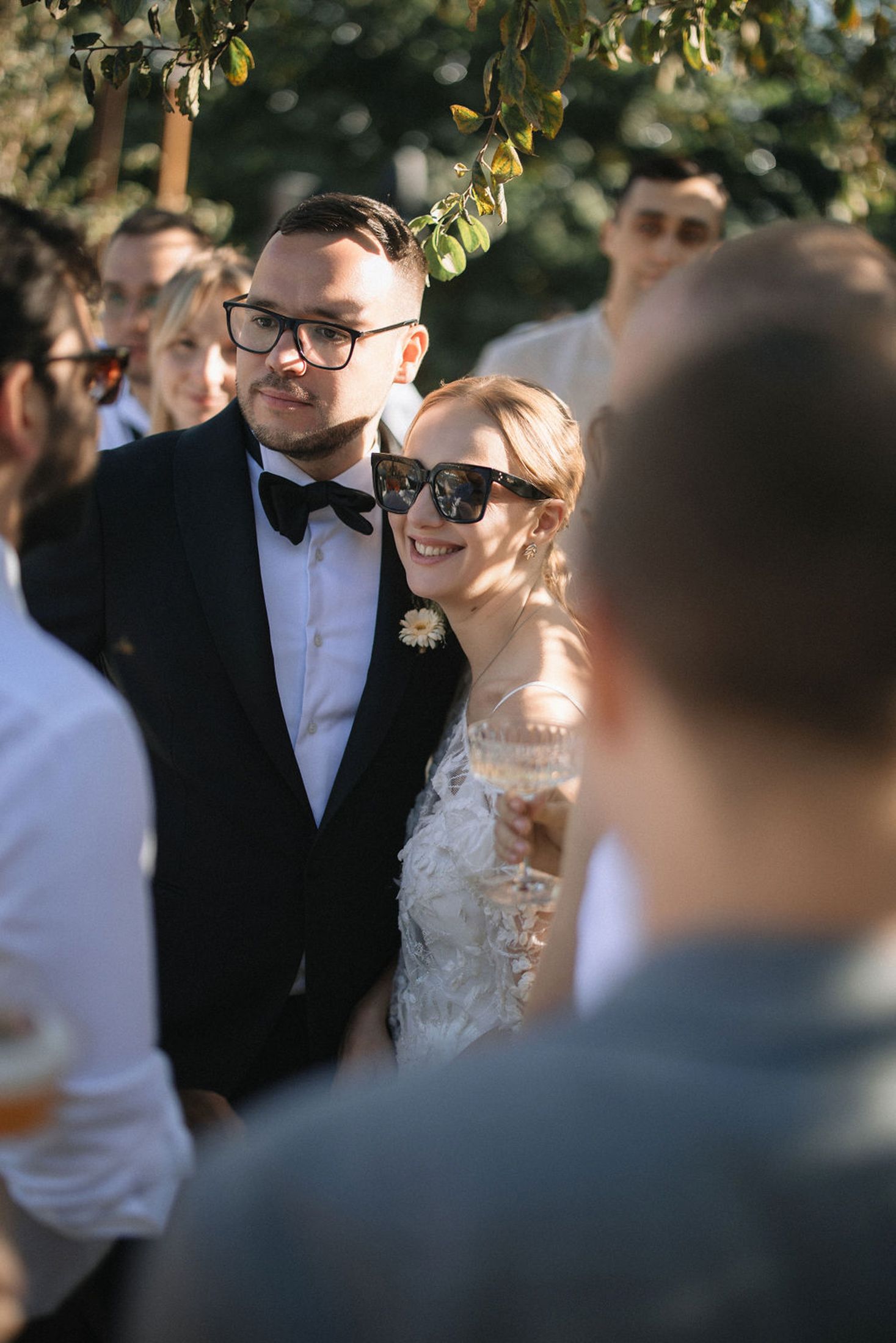 A couple in formal wedding attire share a joyful moment outdoors under sunlit trees.