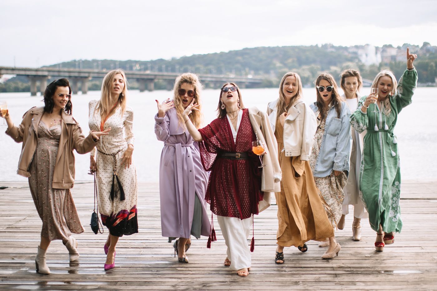 A group wearing coordinated neutral and pastel dresses walking together on a wooden dock by the water.