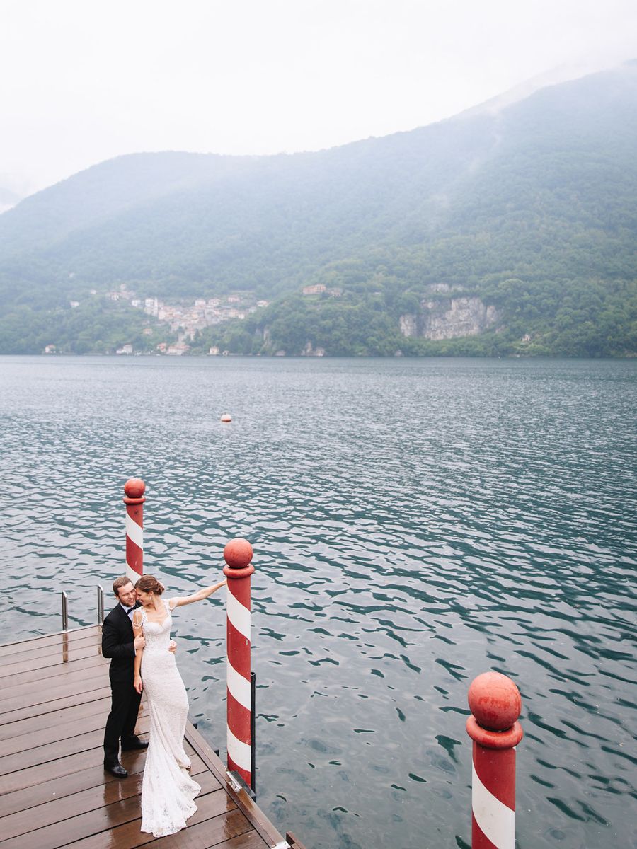 A couple stands on a wooden dock overlooking a misty mountain lake with red and white striped poles in the foreground.