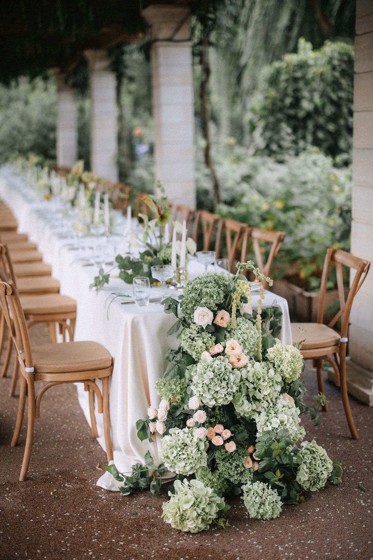 Elegant wedding table setting with cascading hydrangeas and roses on white linen alongside wooden crossback chairs.