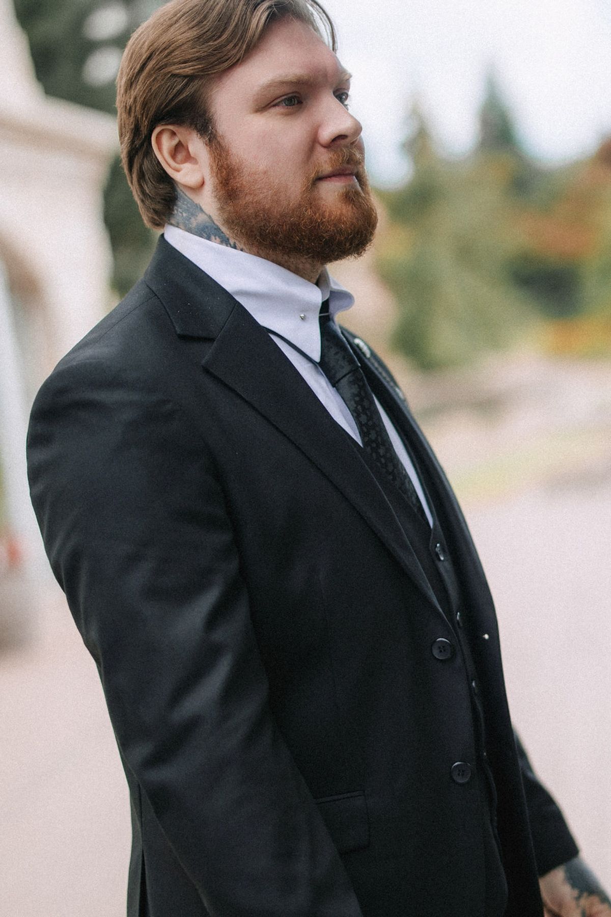 A person in a black suit and tie looking to the side in an outdoor setting.