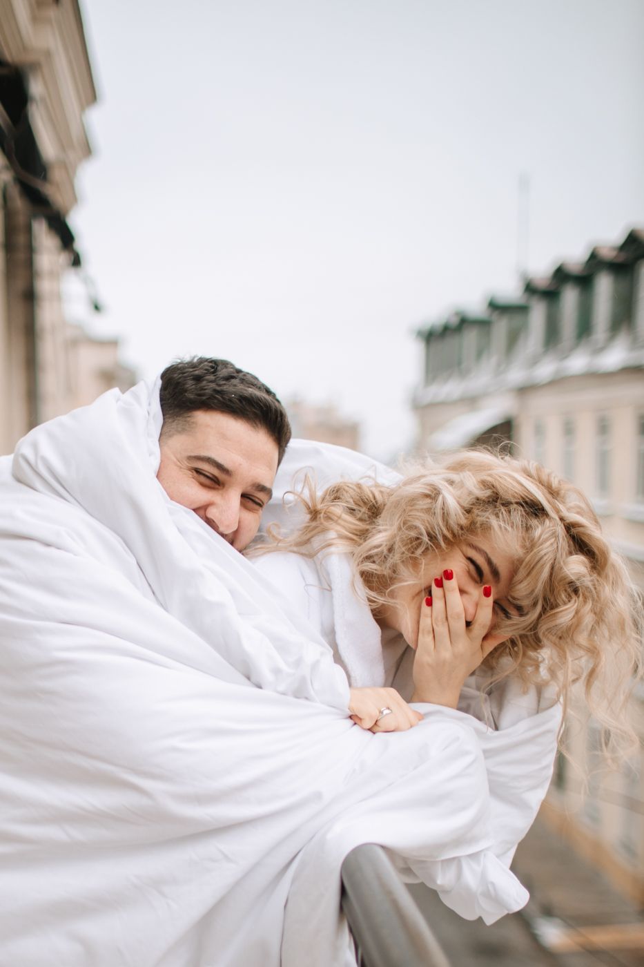 Romantic moment shared between couple wrapped in white blankets on city balcony.