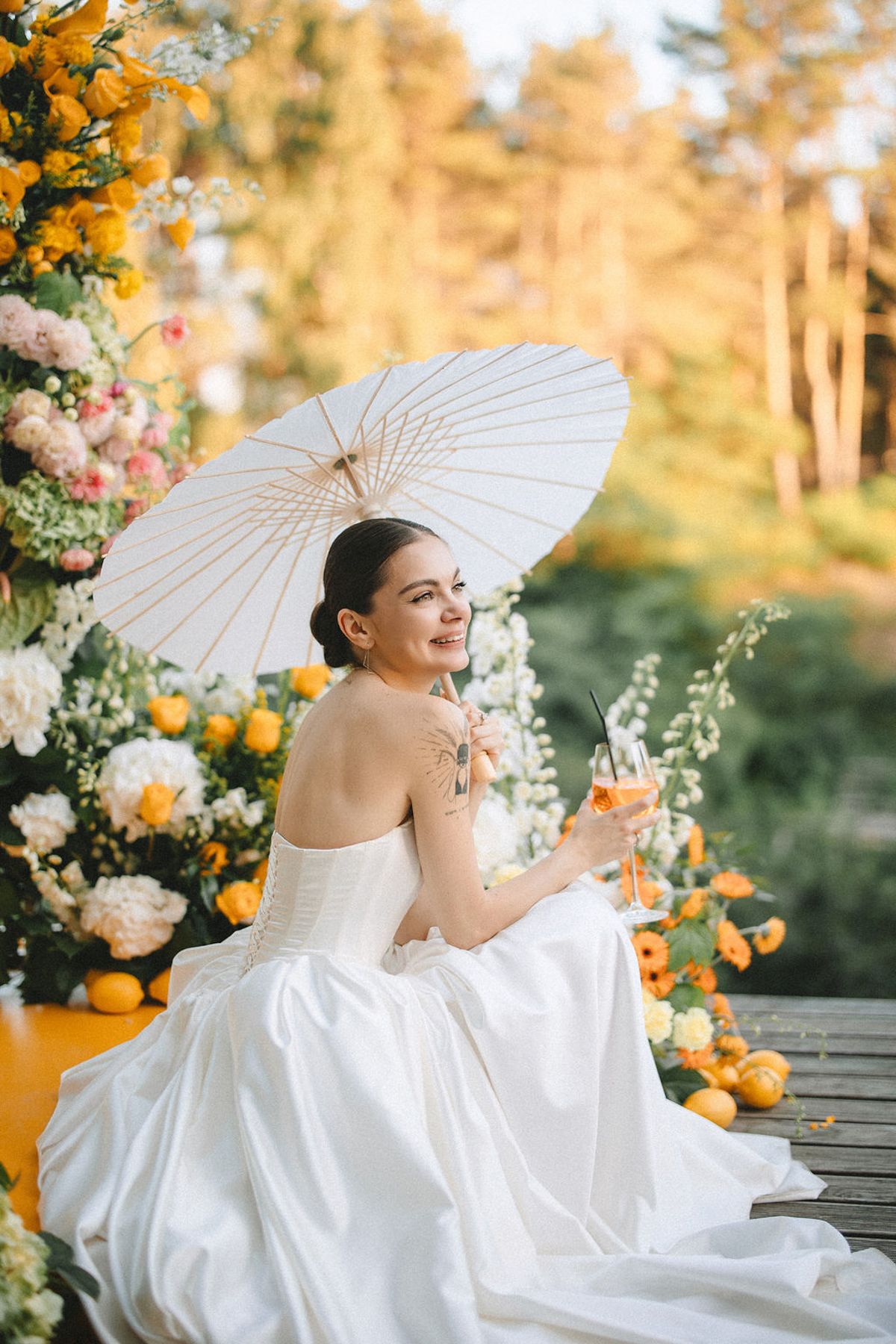A bride in a white wedding gown poses gracefully with a parasol among citrus and floral decorations at sunset.